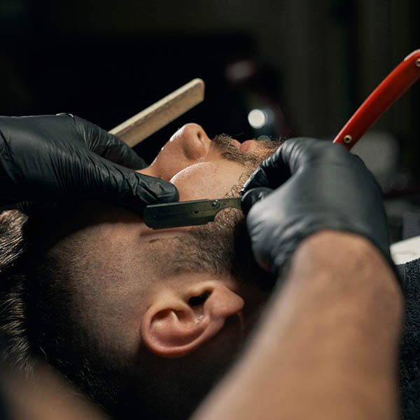 Close up of barbers hands in black gloves shaving bearded client with sharp red razor. Handsome bearded man is getting shaved by hairdresser at the barbershop.Concept of professional customer service