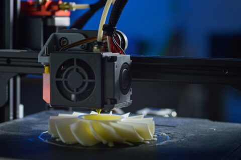Close-up of a 3D printer extruder depositing melted filament to form a layered, spiral-shaped plastic object on a build plate.