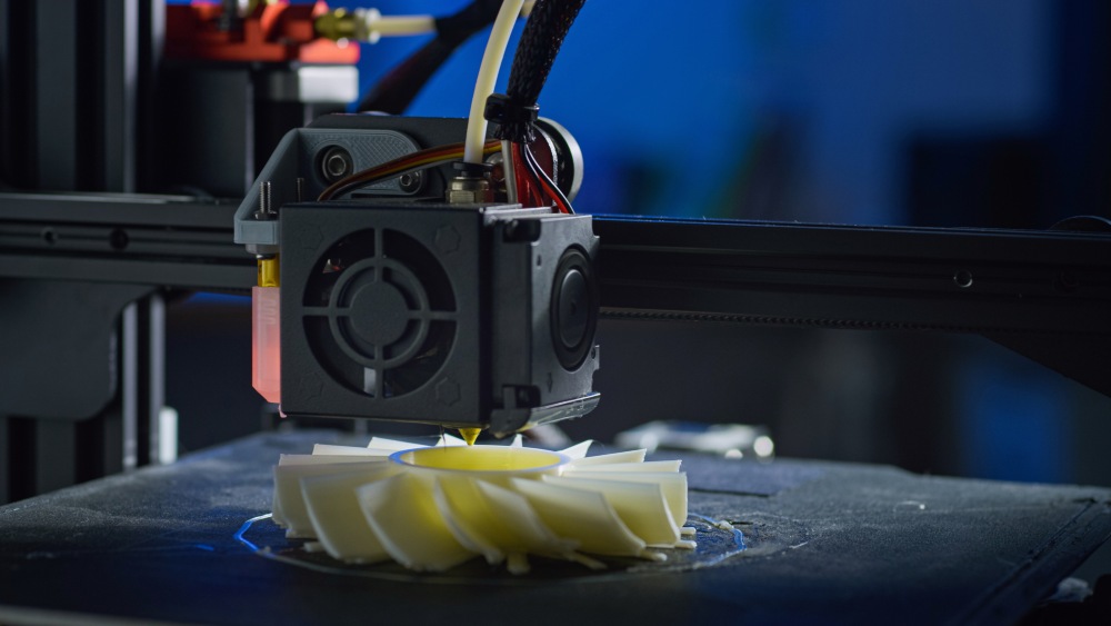 Close-up of a 3D printer extruder depositing melted filament to form a layered, spiral-shaped plastic object on a build plate.